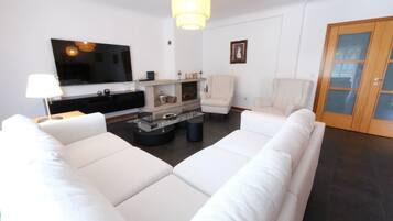 A modern living room with a large flat-screen TV, white sofas, a glass coffee table, and a fireplace.
