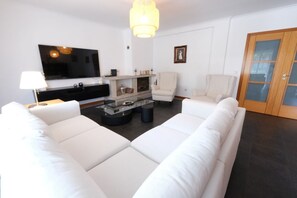 A modern living room with a large flat-screen TV, white sofas, a glass coffee table, and a fireplace.