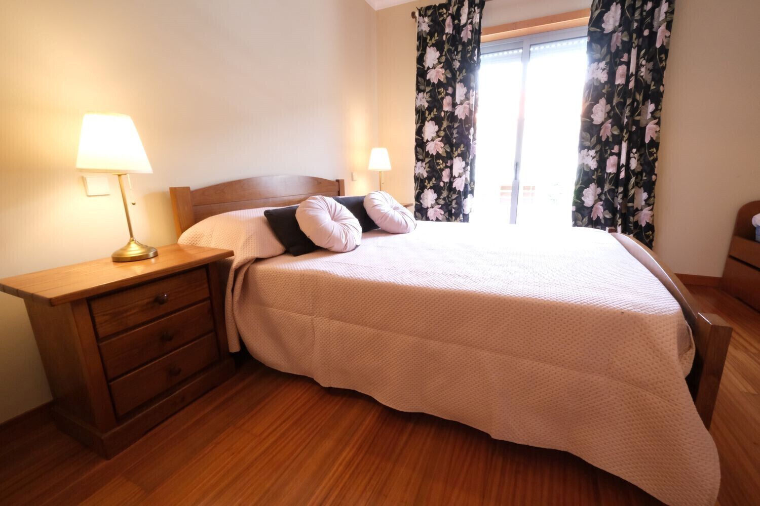A neatly made bed with a headboard, two bedside lamps, a wooden nightstand with drawers, and a window with floral curtains.