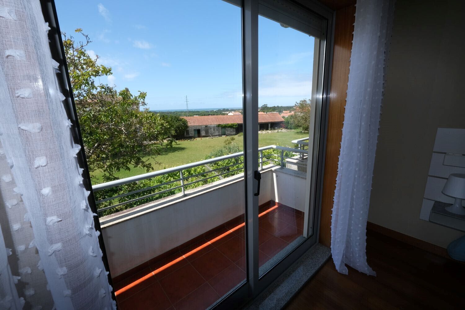 A balcony with a glass door, a view of a green landscape, and a white lamp on the wall.