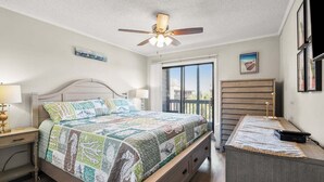 Un dormitorio con cama, mesitas de noche, un escritorio, ventilador de techo y vistas a través de un ventanal.