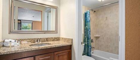 A bathroom with a granite countertop, wooden cabinets, a large mirror, a toilet, and a shower with a blue curtain.