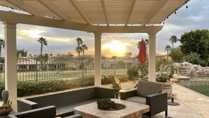 A covered outdoor seating area with wicker furniture, a stone table, and a view of a pool and landscape.