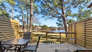 A wooden deck with a table and chairs, overlooking a lake with houses in the background. - Book 3 Nights & Get 1 Night Free* Charming Cottage*6 Seat Cart*Lake*Beach & Pool (Miramar Beach)