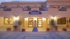 Ein Hotelzugang mit einem Schild, auf dem 'Dolcestate' steht, mit einer Steinfassade, Topfpflanzen und einem blauen Teppich, der zu den Türen führt.