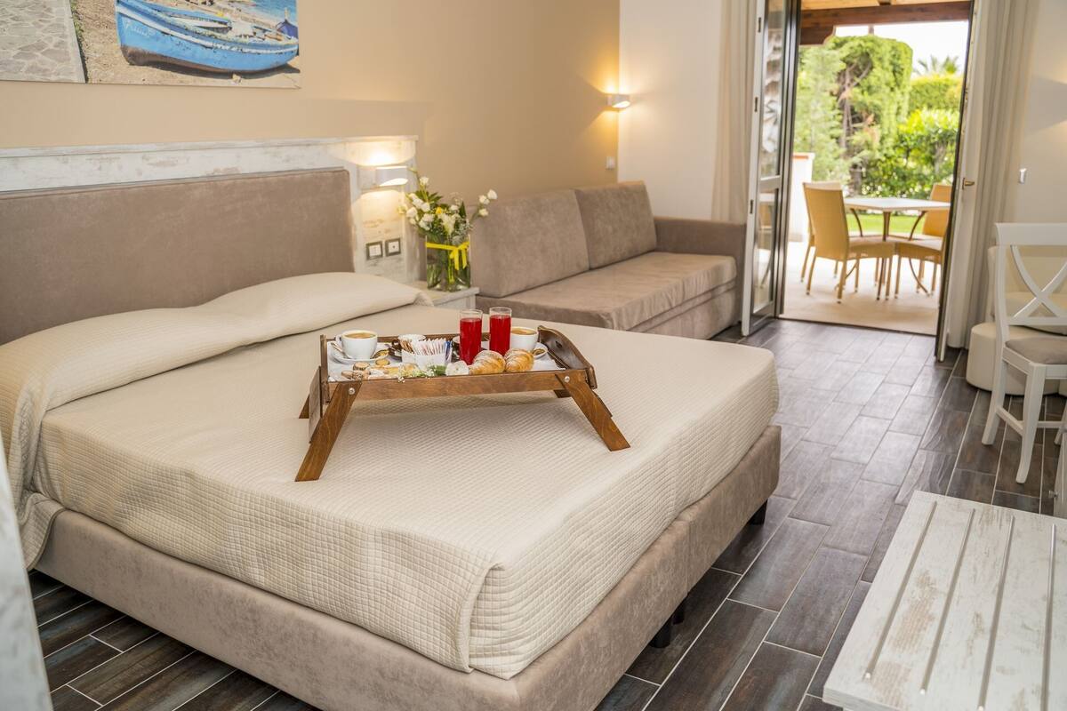 A hotel room with a bed, a wooden tray with breakfast items, a sofa, and a view of the outdoors.