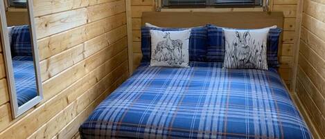 A wooden cabin bedroom with a bed, two pillows with animal prints, a window with blinds, and a mirror on the wall.
