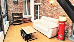 A cozy living room with a brick wall, a white sofa, a wooden coffee table, and a red gas cylinder. - Le Greyhound - Grand Appartement Centre-ville (Soissons)