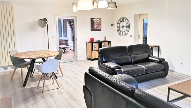 A modern living room with a black leather sofa, a wooden dining table with chairs, and a large wall clock. - Le Greyhound - Grand Appartement Centre-ville (Soissons)