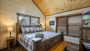 Une chambre confortable avec un plafond en bois, un lit recouvert d’une couette à carreaux, un banc et une vue sur une maison par la fenêtre.