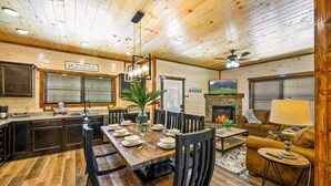 L’intérieur chaleureux d’un chalet comprend une table à manger en bois, préparée pour quatre personnes, une cheminée, un coin salon avec un canapé et une cuisine dotée de meubles sombres.