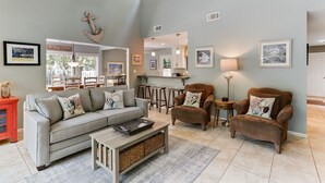 A spacious living room with a grey sofa, two brown armchairs, a coffee table, and a kitchen in the background.