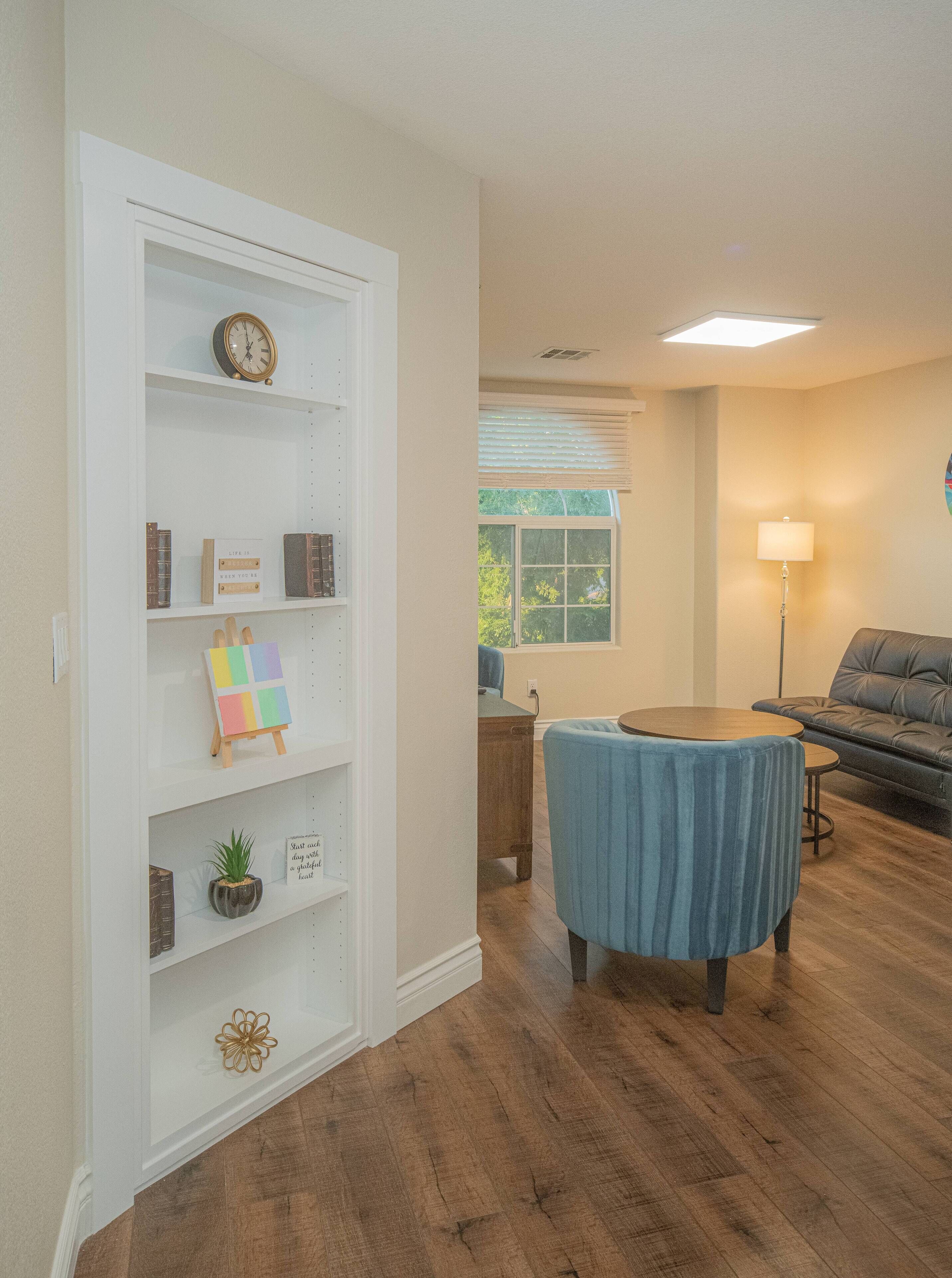 A living room with a wooden floor, a blue chair, a sofa, a bookshelf, and a window with blinds.