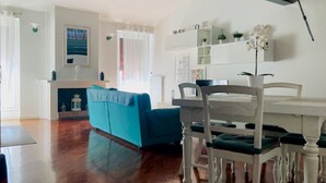 Amplia sala de estar con un sofá azul, una mesa de comedor blanca y sillas. Se ve una chimenea al fondo, y hay estantes con plantas y objetos decorativos.