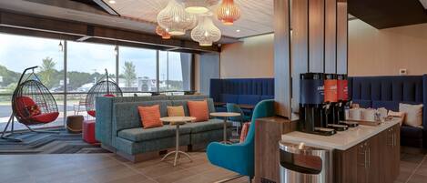 A modern hotel lobby with seating areas, a coffee station, and large windows offering a view of the parking lot.