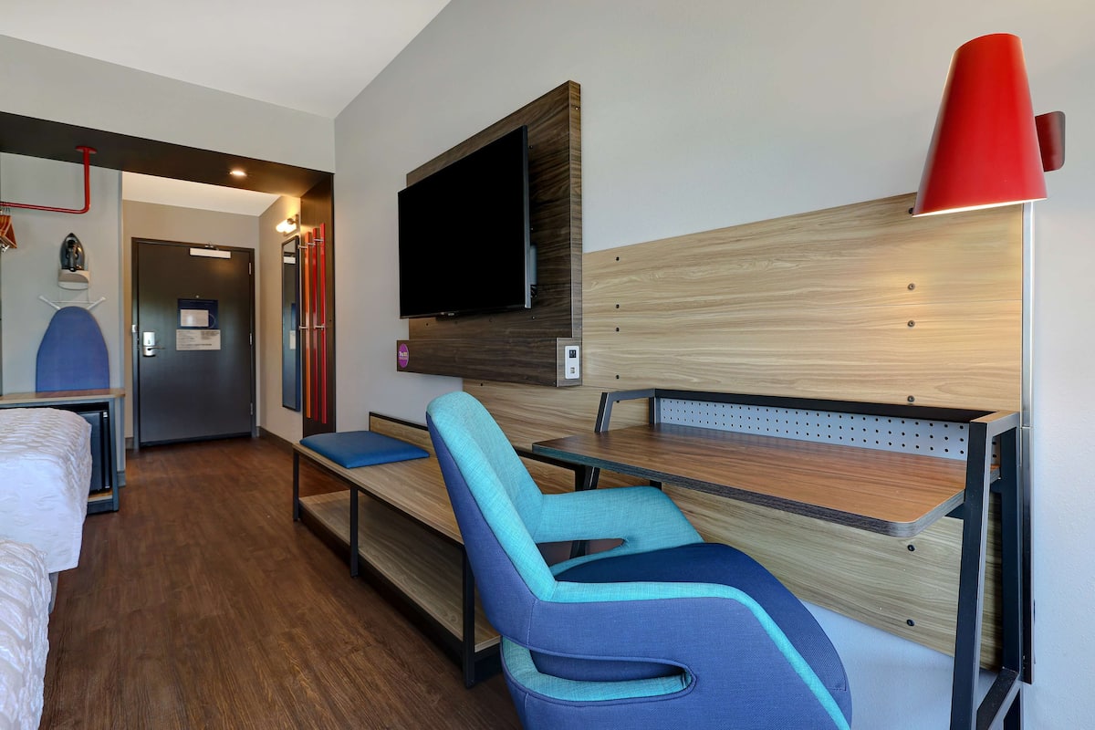A hotel room with a bed, a desk with a chair, a television mounted on the wall, and a red lamp.