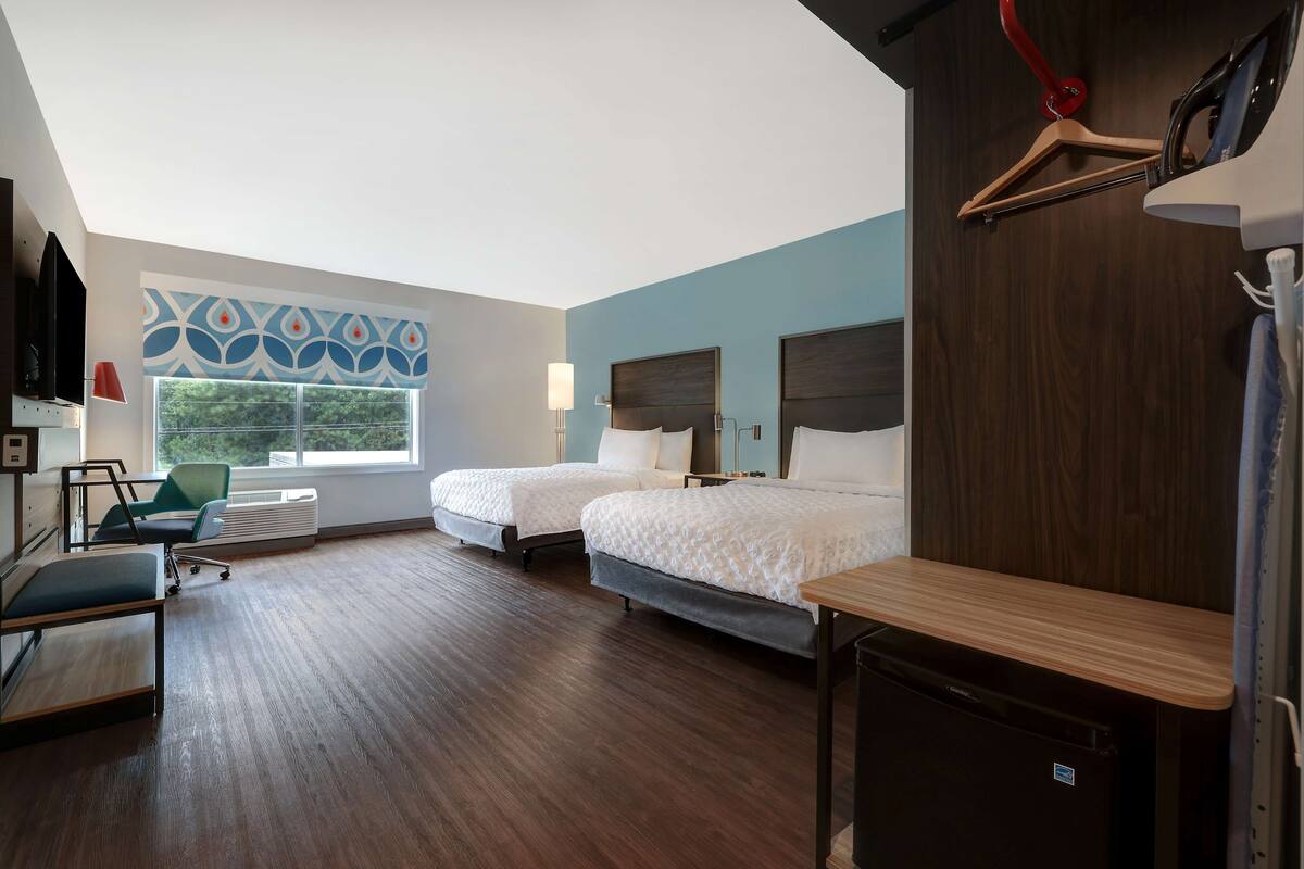 A hotel room with two beds, a desk, a chair, a TV, and a window with a view of greenery.