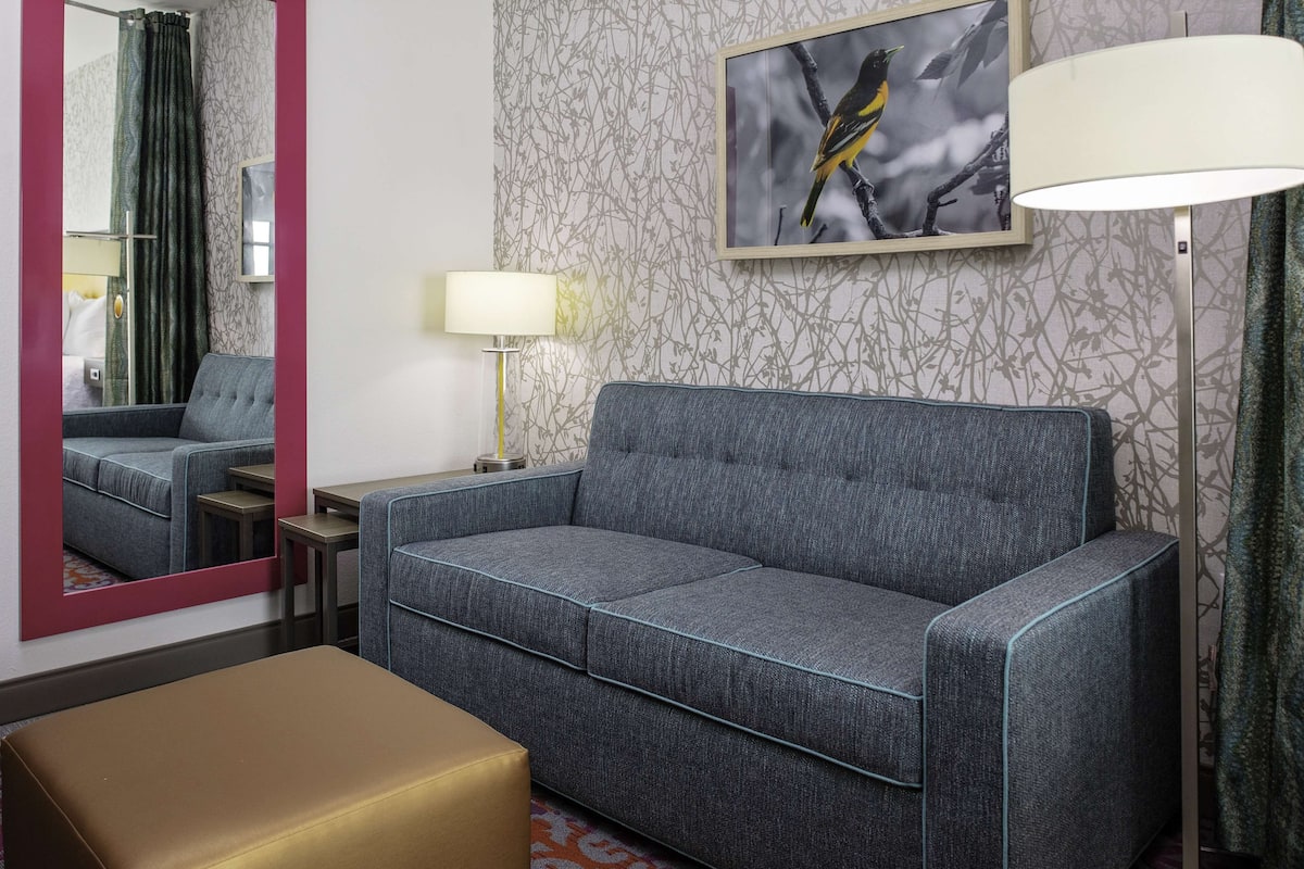 A hotel room with a grey sofa, a small table, a lamp, a mirror, and a framed picture of a bird on the wall.