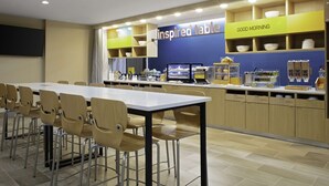 A modern kitchen area with a long dining table and chairs, a TV mounted on the wall, and various kitchen appliances and utensils. - Home2 Suites by Hilton Baton Rouge Citiplace (Baton Rouge)