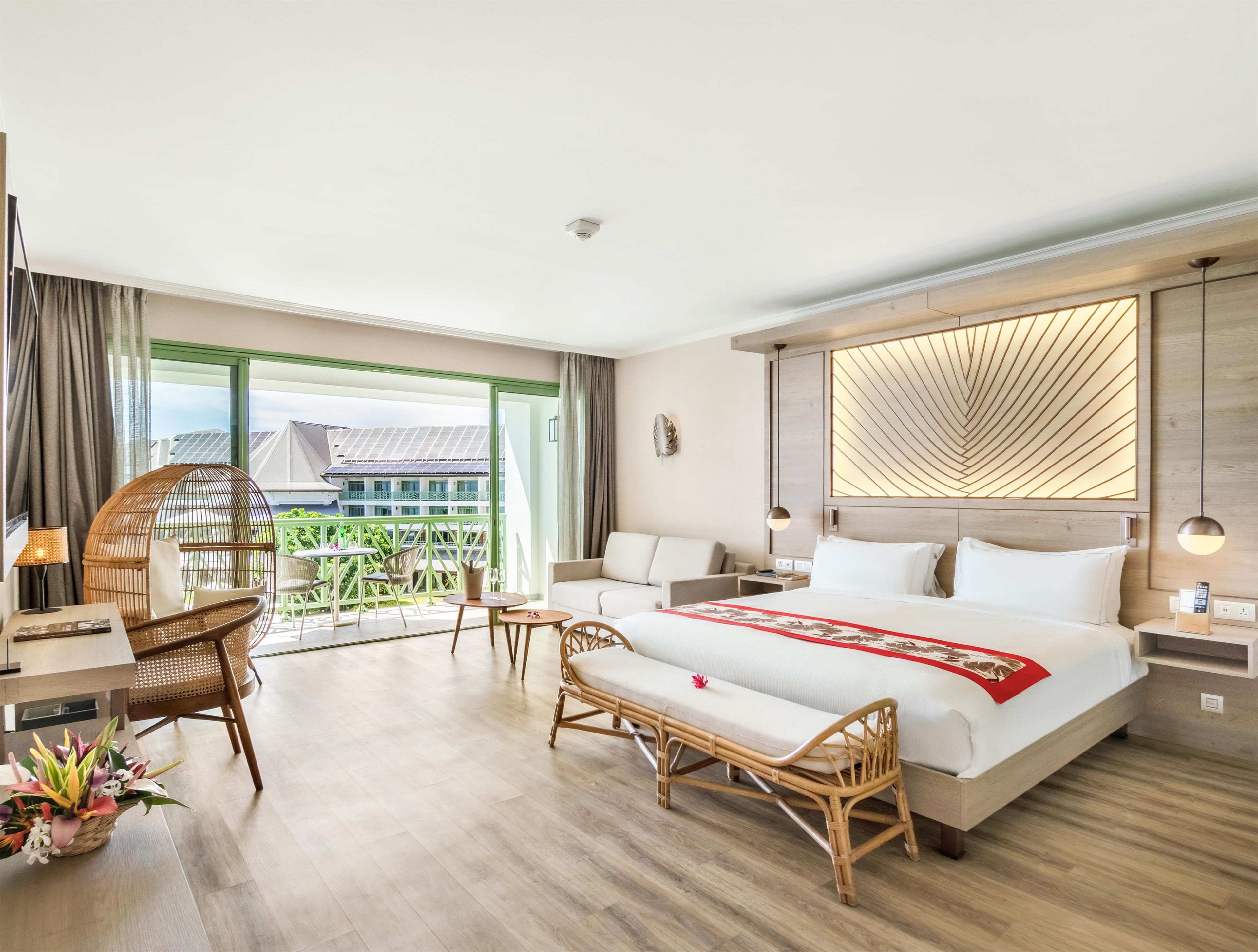 A hotel room with a bed, a sofa, a wicker chair, a small table, and a balcony with a view of buildings.