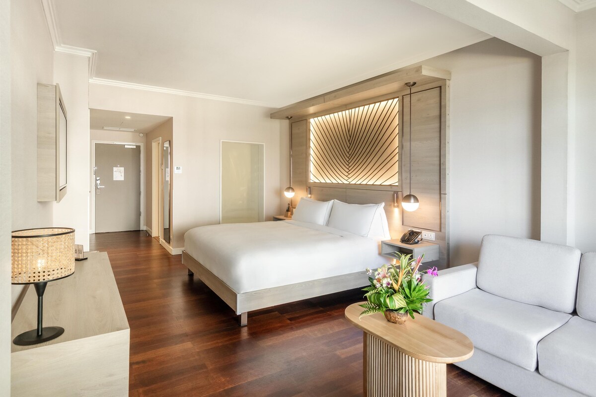 Quarto de hotel com uma cama grande, um sofá, uma escrivaninha e um painel decorativo na parede.
