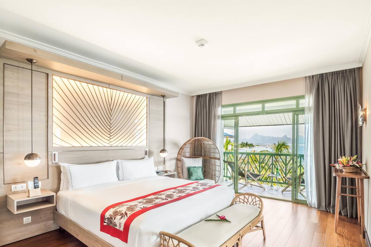 A modern hotel room with a large bed, a balcony with a view, and a wooden floor.