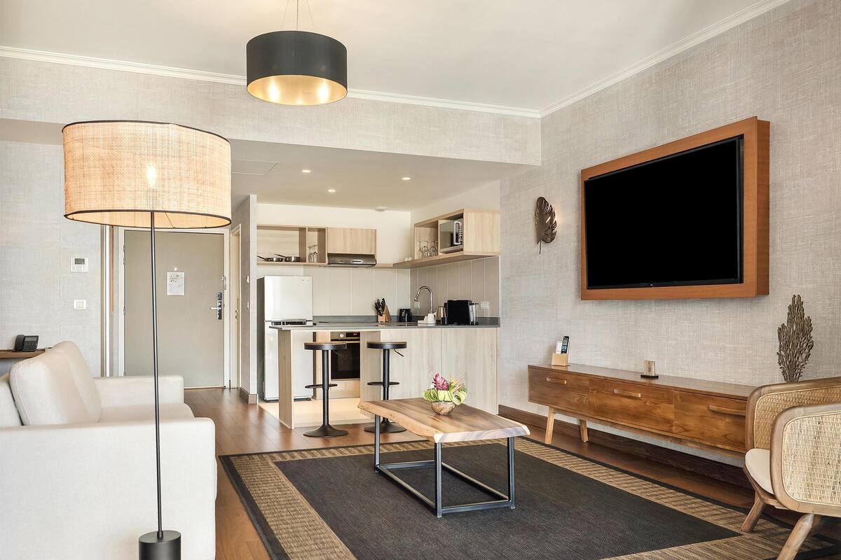 A modern living room with a sofa, a coffee table, a TV, and a kitchen area in the background.