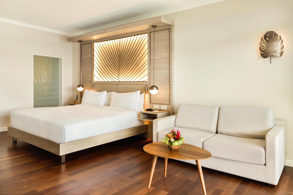 Quarto de hotel com uma cama, um sofá, uma mesinha com um vaso de flores, luminária na parede e uma janela grande com padrão geométrico.