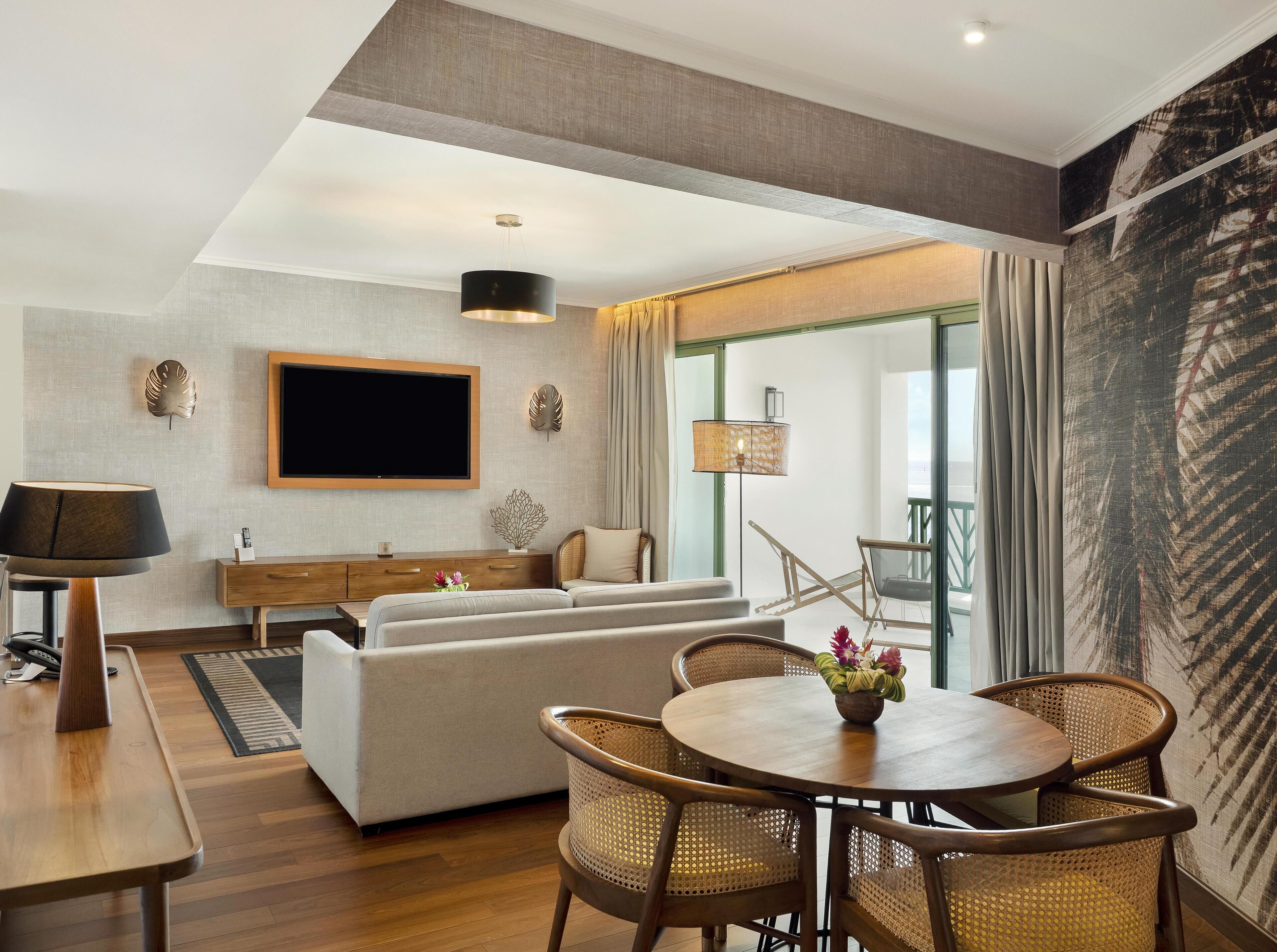 Un salon moderne comprenant un canapé, une table à manger et des chaises, un téléviseur à écran plat fixé au mur et une grande fenêtre avec des rideaux.