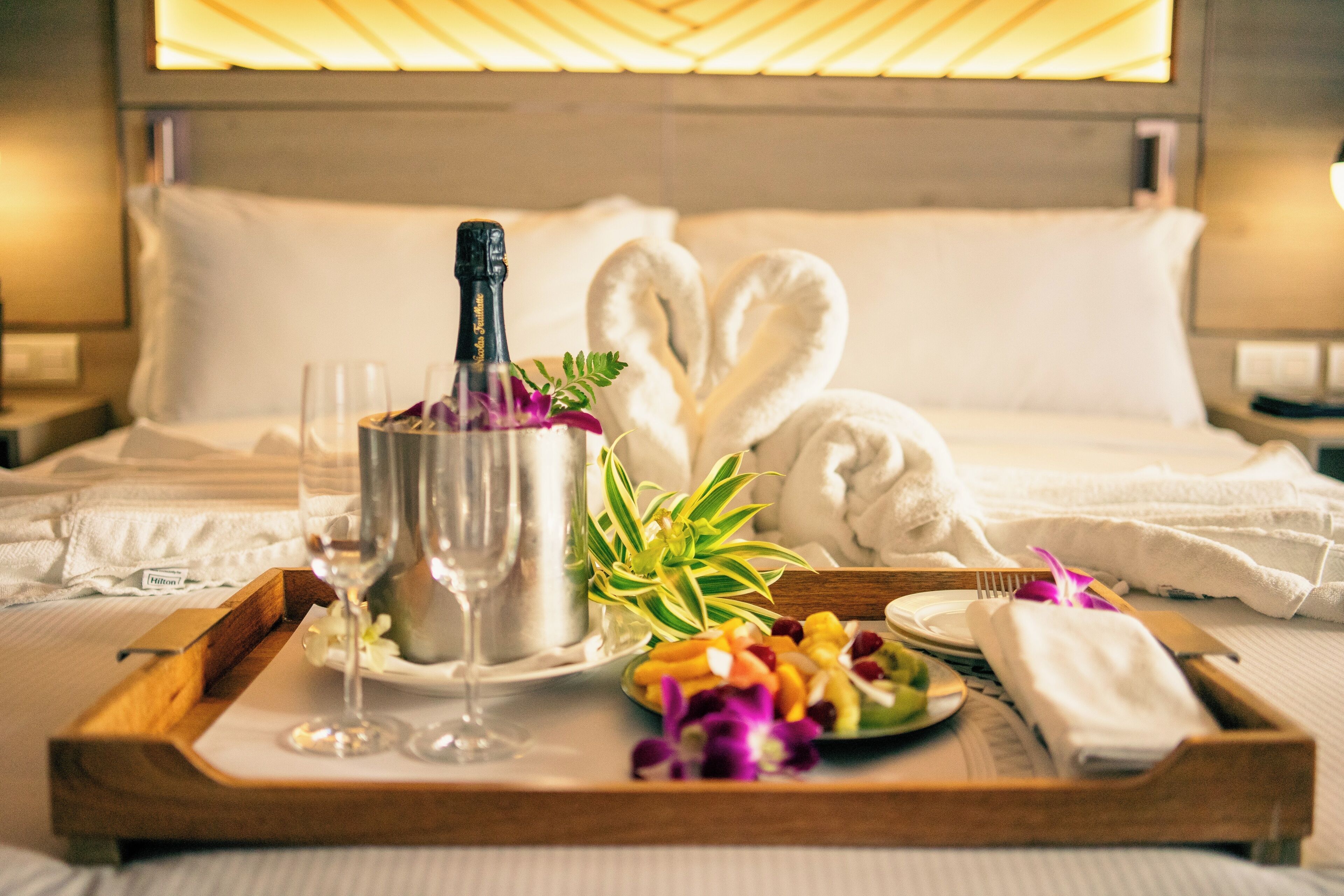 A neatly arranged breakfast tray on a bed with a bottle of champagne, glasses, and a plate of fruit.