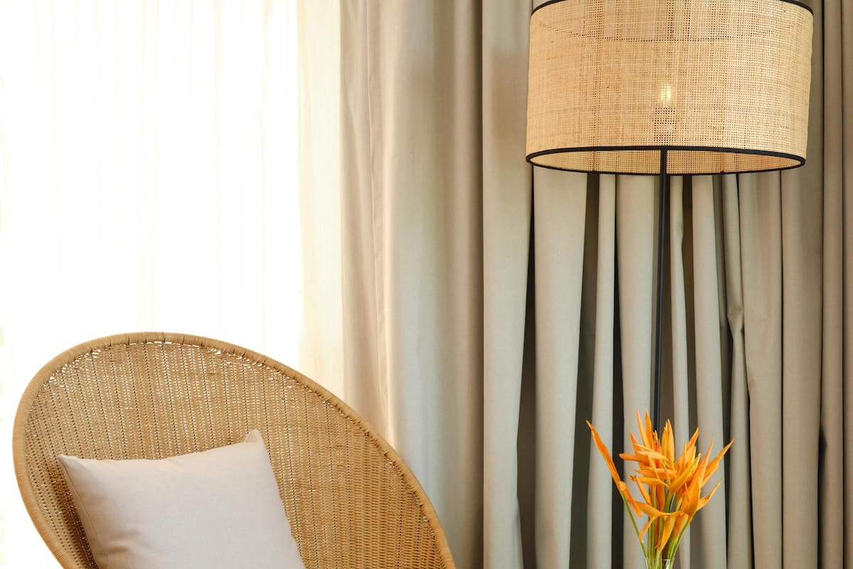 A wicker chair with a white cushion, a round wooden side table with a vase of orange flowers, and a large woven floor lamp.