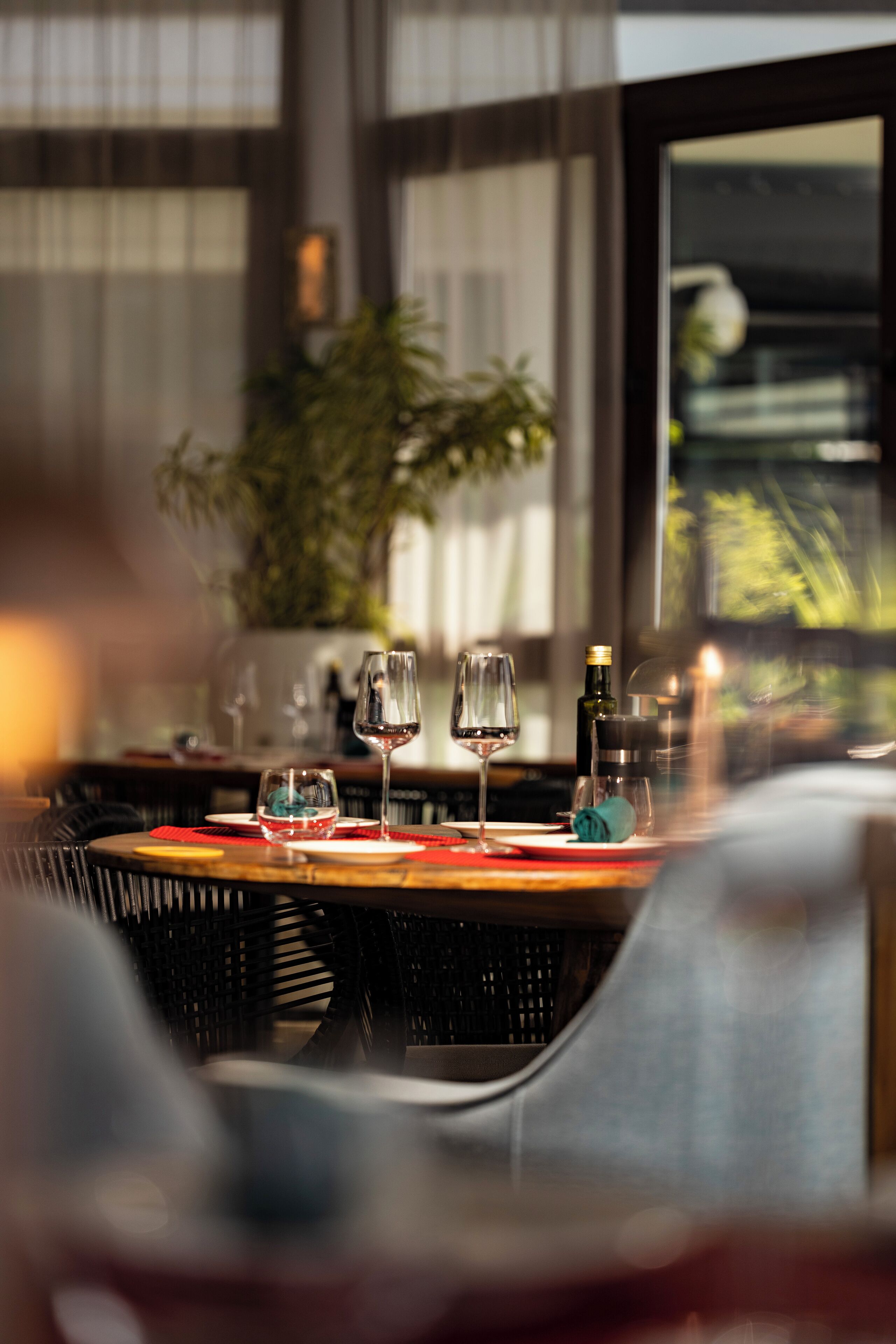 Een eettafel met wijnglazen en een fles, geplaatst in een restaurant met rieten stoelen en een groot raam op de achtergrond.