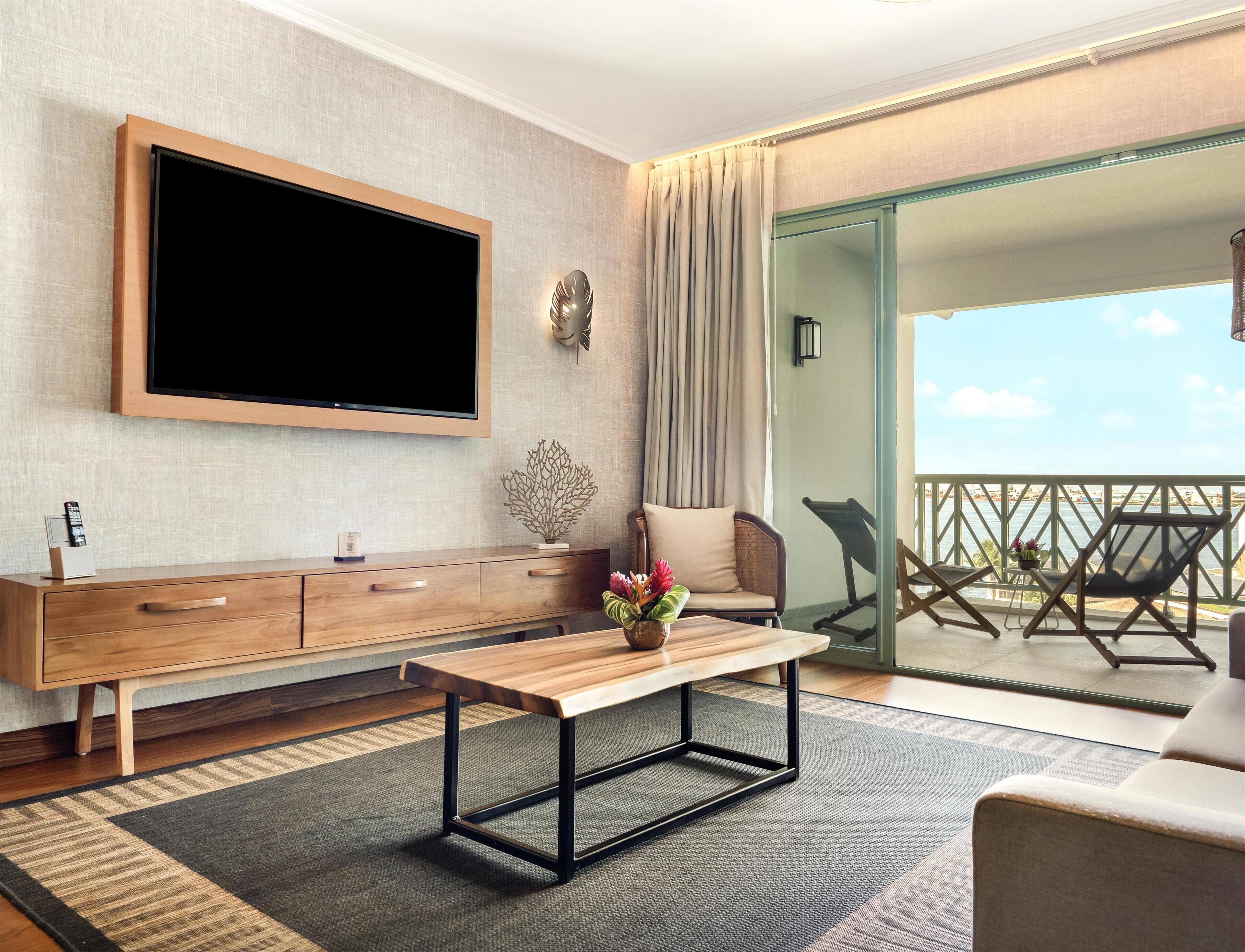 A modern living room with a flat-screen TV, wooden furniture, a bench, and a view of the sea.