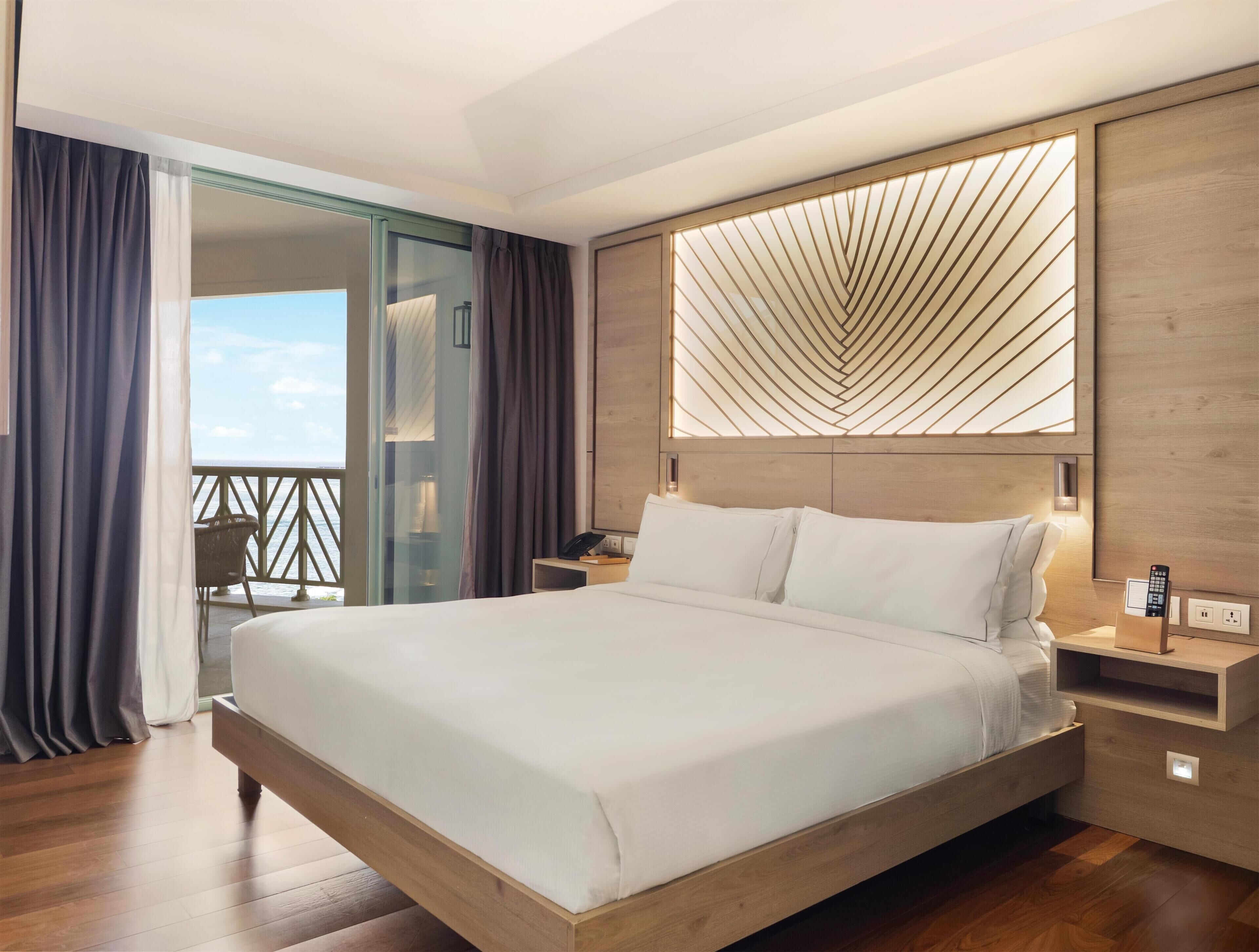 A modern hotel room with a large bed, a glass door leading to a balcony, and a wooden headboard with a geometric light design.