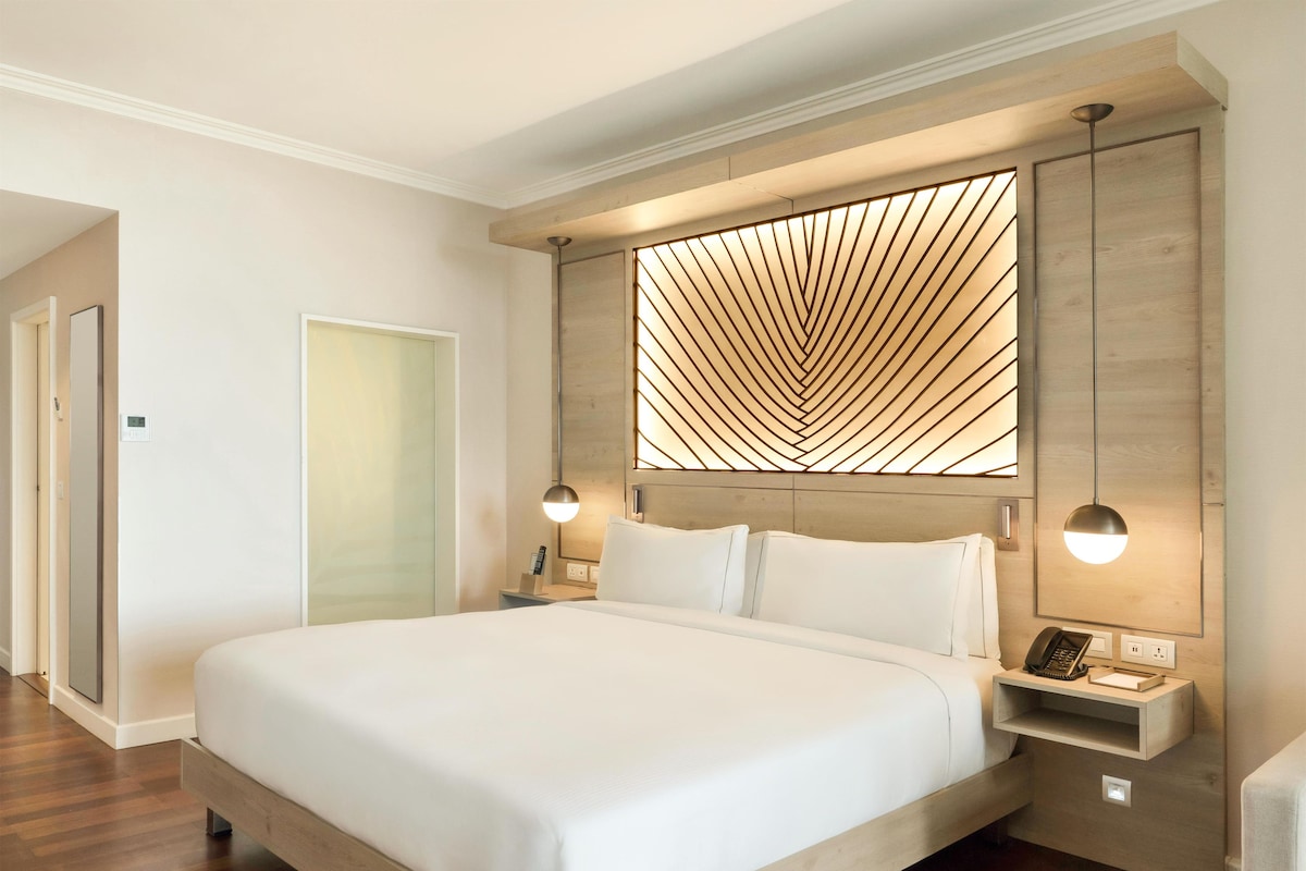 Quarto de hotel moderno com uma cama grande, cabeceira de madeira, luminárias de cabeceira e um painel de luz embutido.