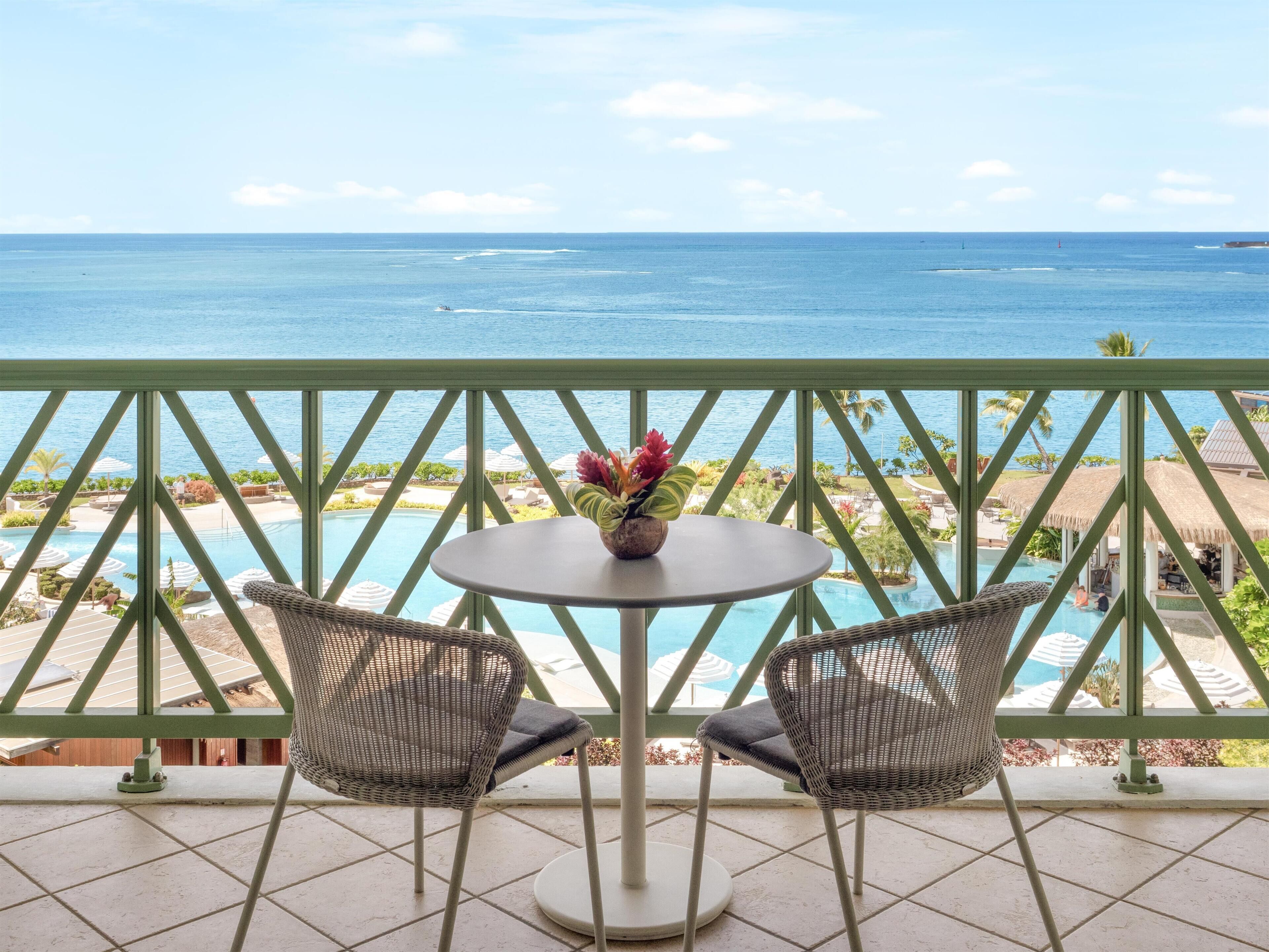 A balcony with a view of the ocean, a pool, and two wicker chairs with a table.