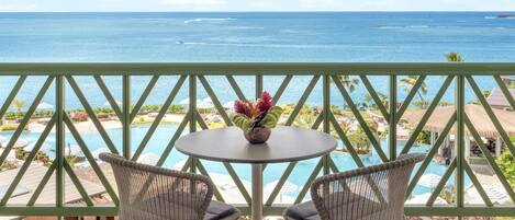 A balcony with a view of the ocean, a pool, and two wicker chairs with a table.