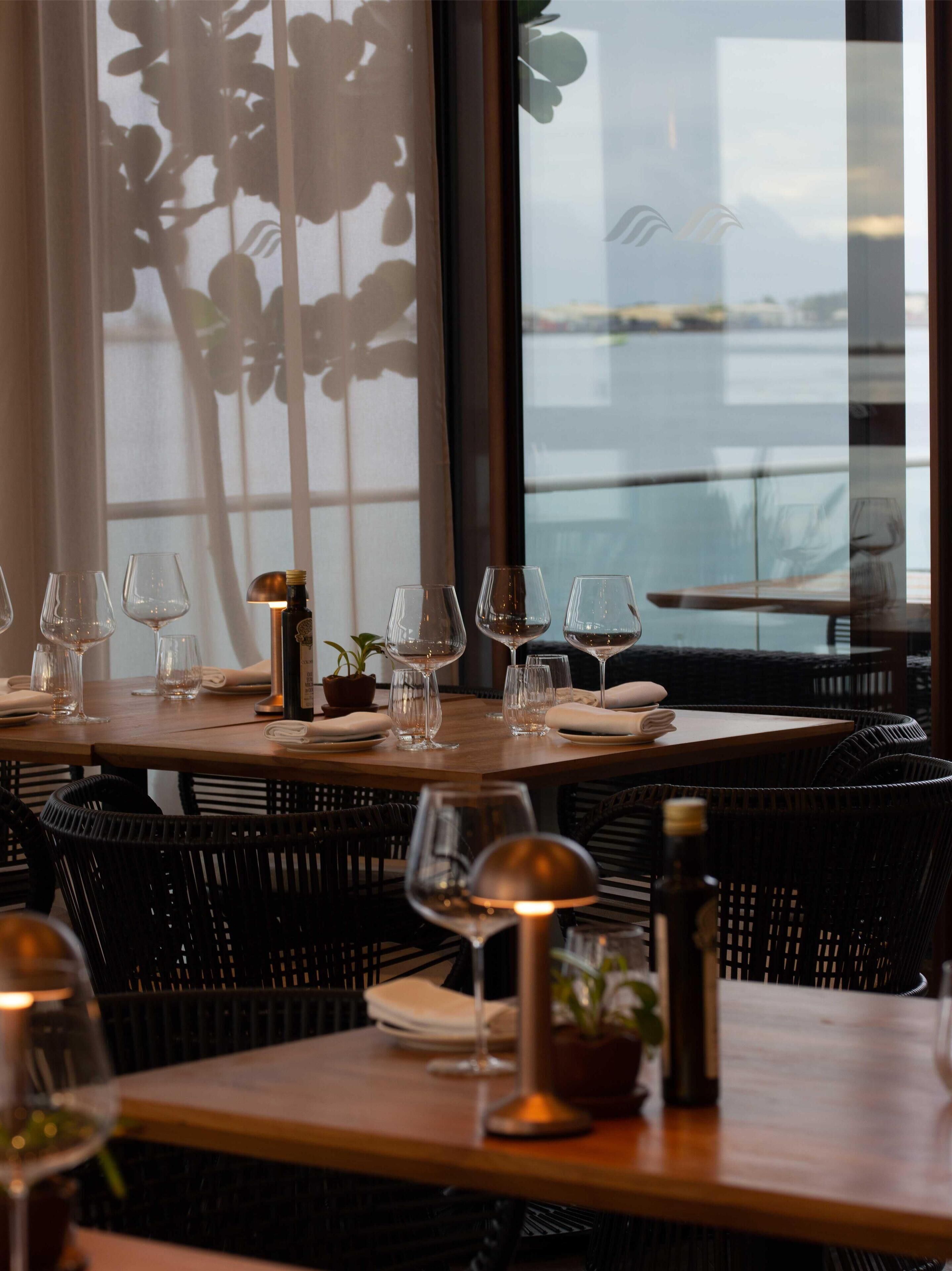 A restaurant with a view of the sea, featuring tables set with wine glasses and candles, and wicker chairs.