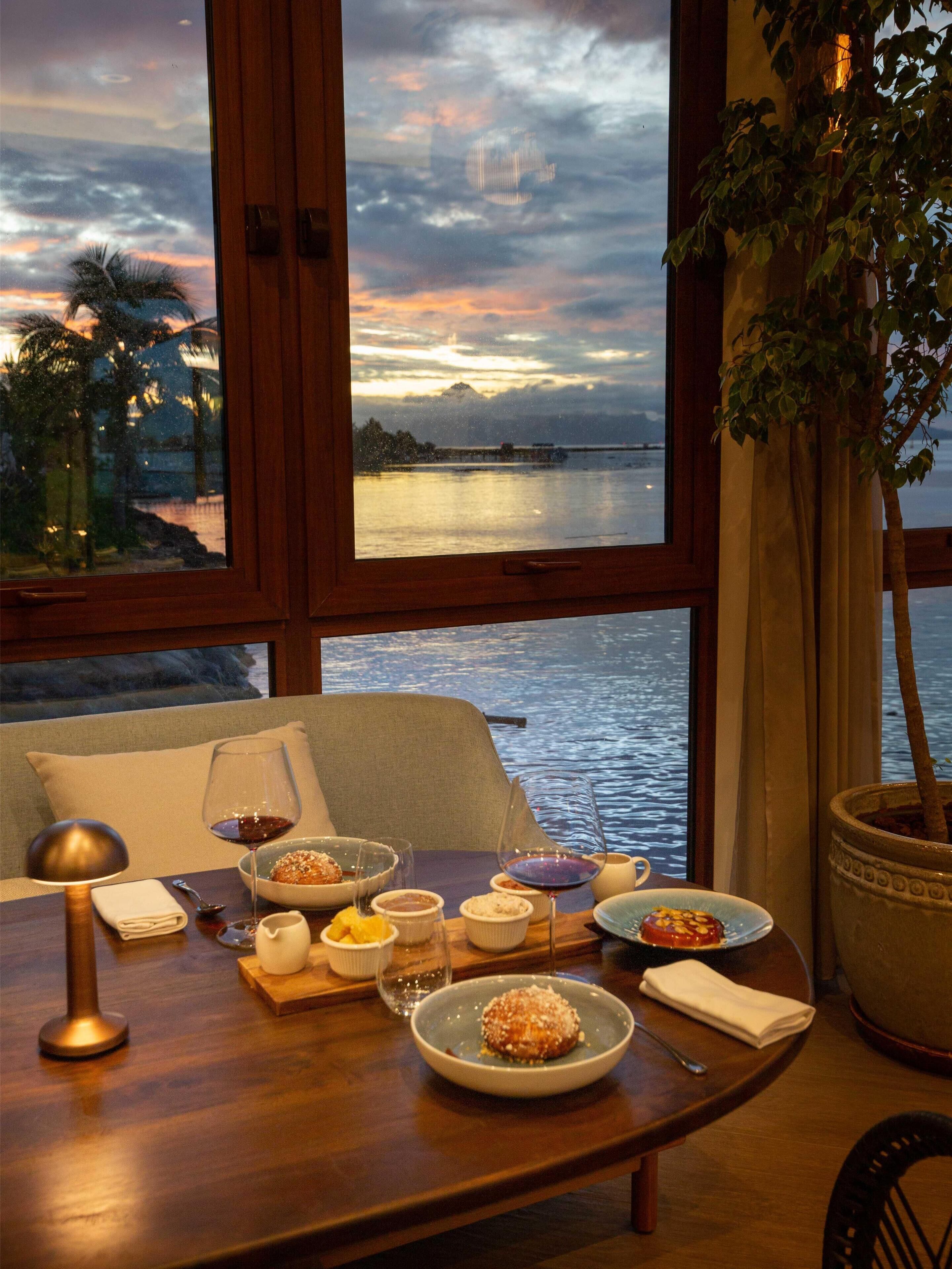Uma mesa de jantar com pratos de comida, copos e uma vela, em frente a uma grande janela que dá para um corpo de água e uma montanha ao pôr do sol.
