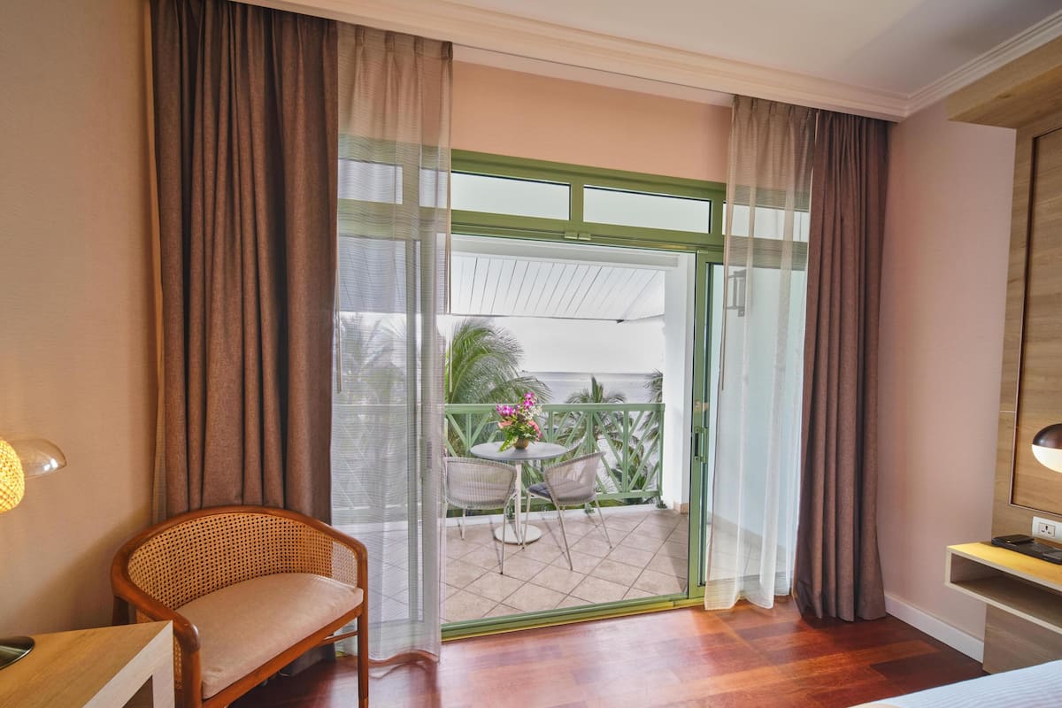 A hotel room with a bed, a wooden chair, a small table with a plant, and a balcony with a view of palm trees and the sea.