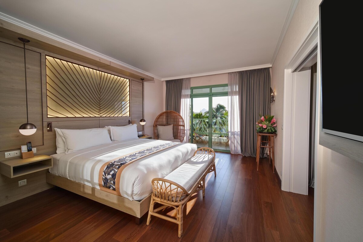 A modern hotel room with a large bed, wooden flooring, a seating area with a chair, a TV, and a balcony with a view of palm trees.
