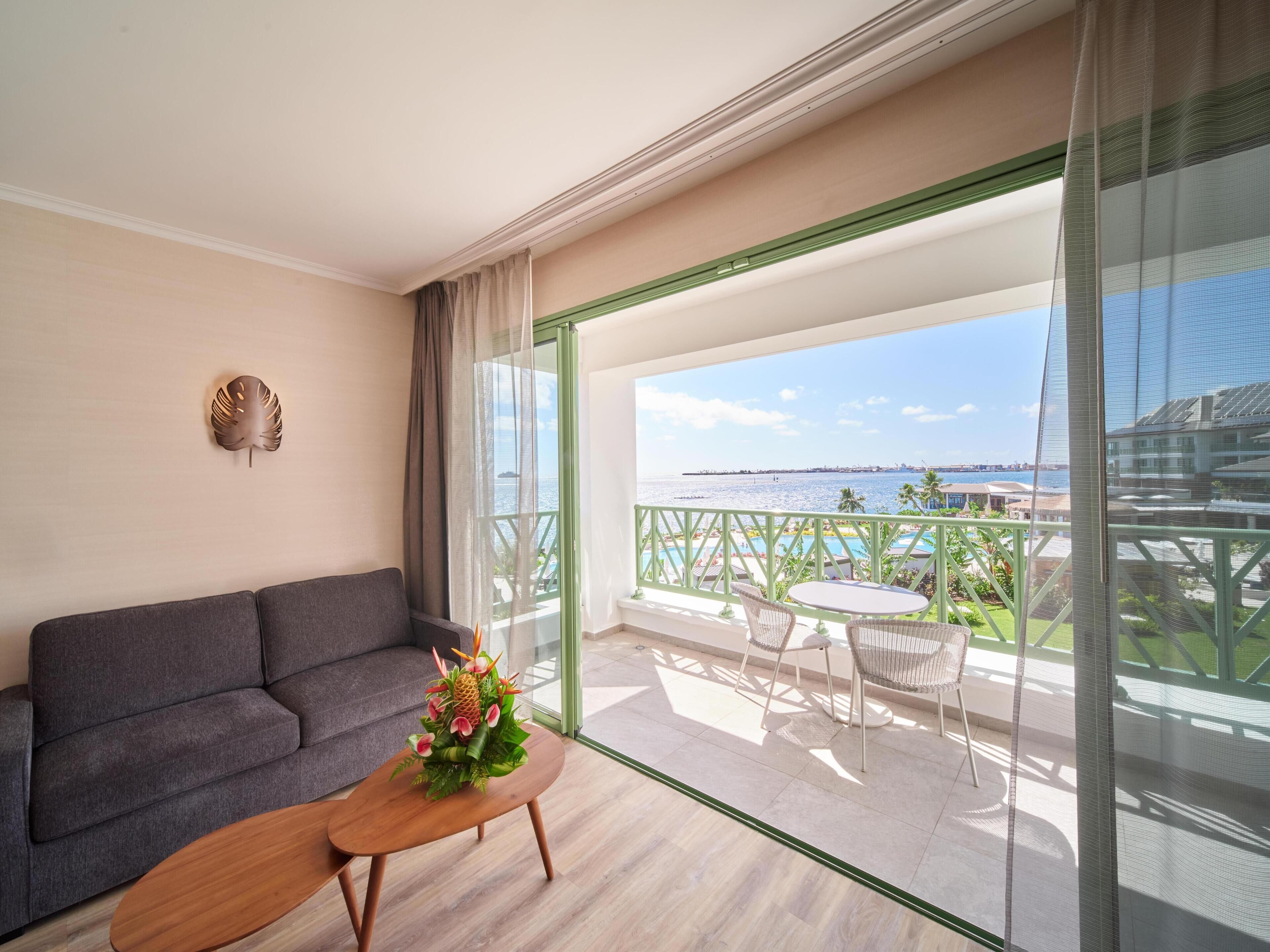 A balcony with a view of the sea, a sofa, a table with a flower arrangement, and a decorative wall piece.