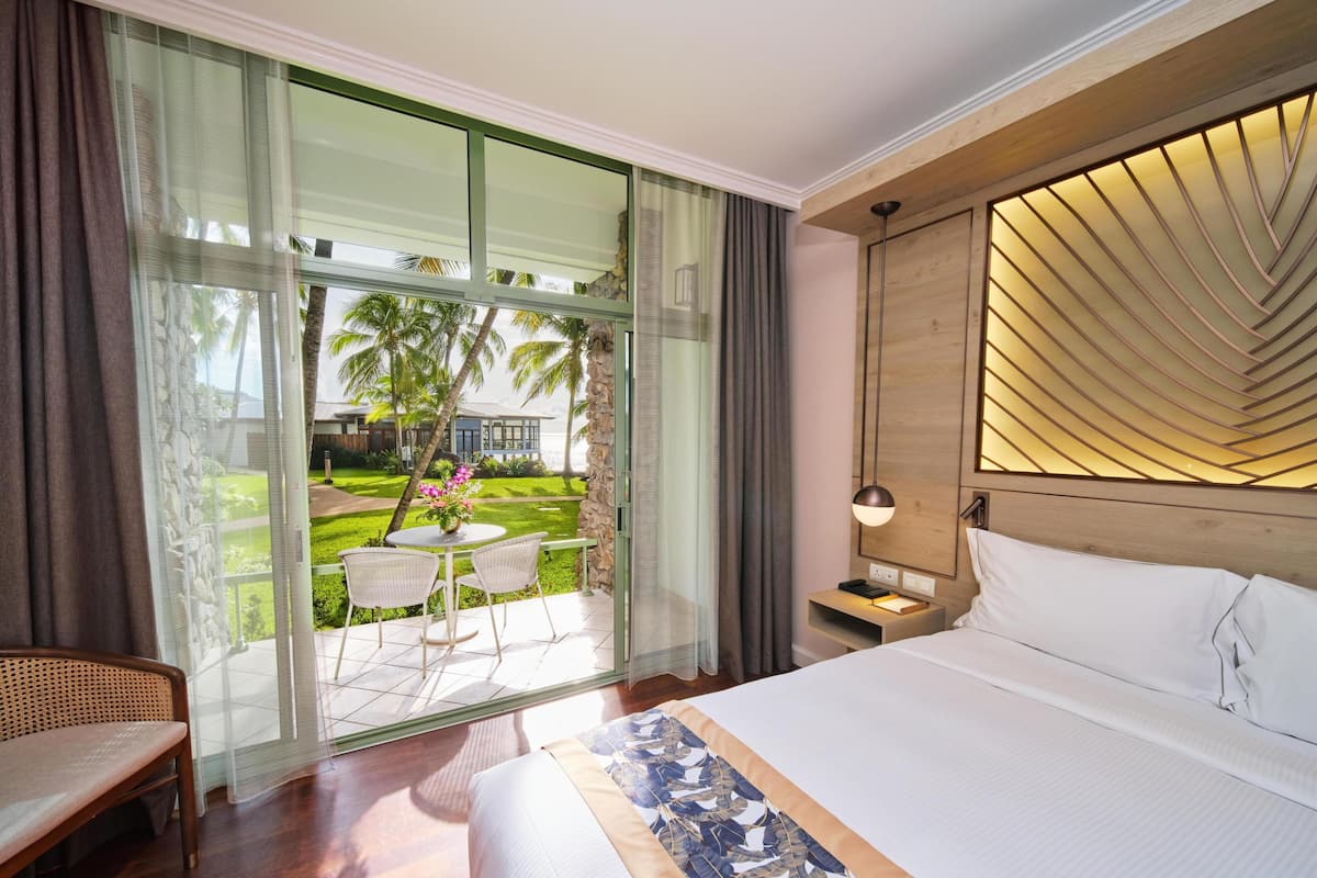A hotel room with a large bed, a bedside table with a lamp, a small table with chairs outside the window, and a view of a garden and palm trees.
