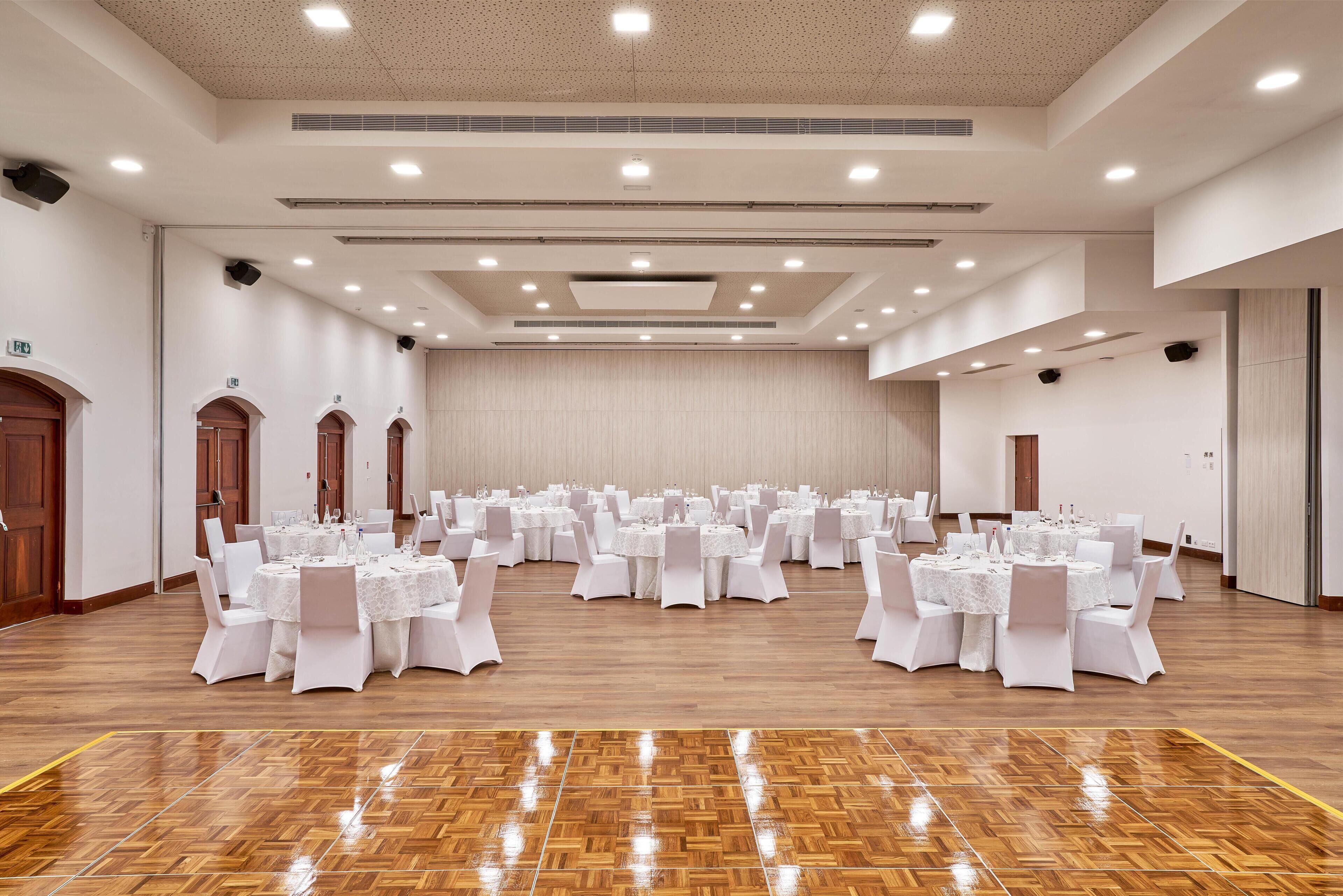 Une grande salle de banquet, bien éclairée, avec de nombreuses tables rondes disposées pour des événements, agrémentée de nappes blanches et de chaises.