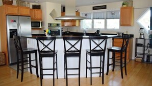 A modern kitchen with wooden cabinets, a central island with bar stools, stainless steel appliances, and a large window providing natural light. - Allowaves- Luxury Town Home with Community Pool & Fishing Pier (Chincoteague)