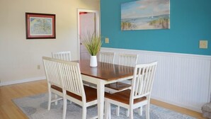 A dining area with a wooden table and white chairs, a blue accent wall, and a framed painting. - Allowaves- Luxury Town Home with Community Pool & Fishing Pier (Chincoteague)