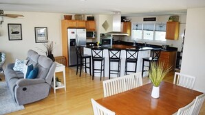 A modern kitchen with a central island, wooden cabinets, and a dining area with a wooden table and white chairs. - Allowaves- Luxury Town Home with Community Pool & Fishing Pier (Chincoteague)