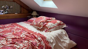 Un dormitorio con techo inclinado, tragaluz, cama con ropa de cama estampada, cabecero de madera y mesita de noche con lámpara.