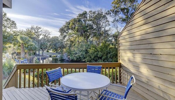 A wooden deck with a round table and blue chairs, surrounded by trees and a clear sky.