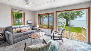 A living room with a grey sofa, a wooden coffee table, and a chair. There is a large window with a view of a lake and a deck outside. - Turquoise Beach Cottage (Luxemburg)